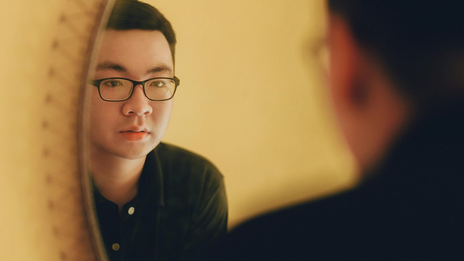 East Asian man with glasses looking at his reflection in a mirror, contemplative mood.