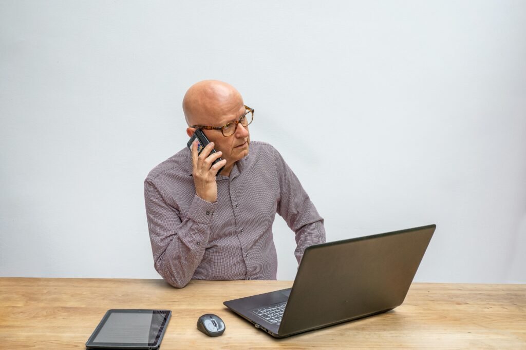 pexels-photo-5511629-5511629 Senior man wearing eyeglasses using cellphone while working on laptop, illustrating modern business communication.