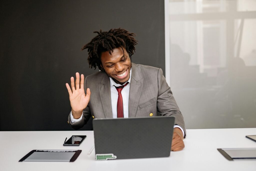 pexels-photo-7792744-7792744 Professional making a video call, waving hand at laptop in modern office setting.