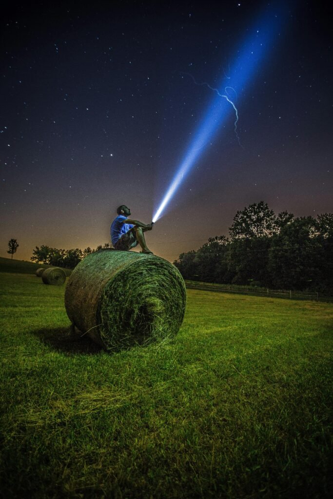 pexels-photo-1816699-1816699 A man sits on a hay bale in a field, shining a flashlight into the night sky full of stars.