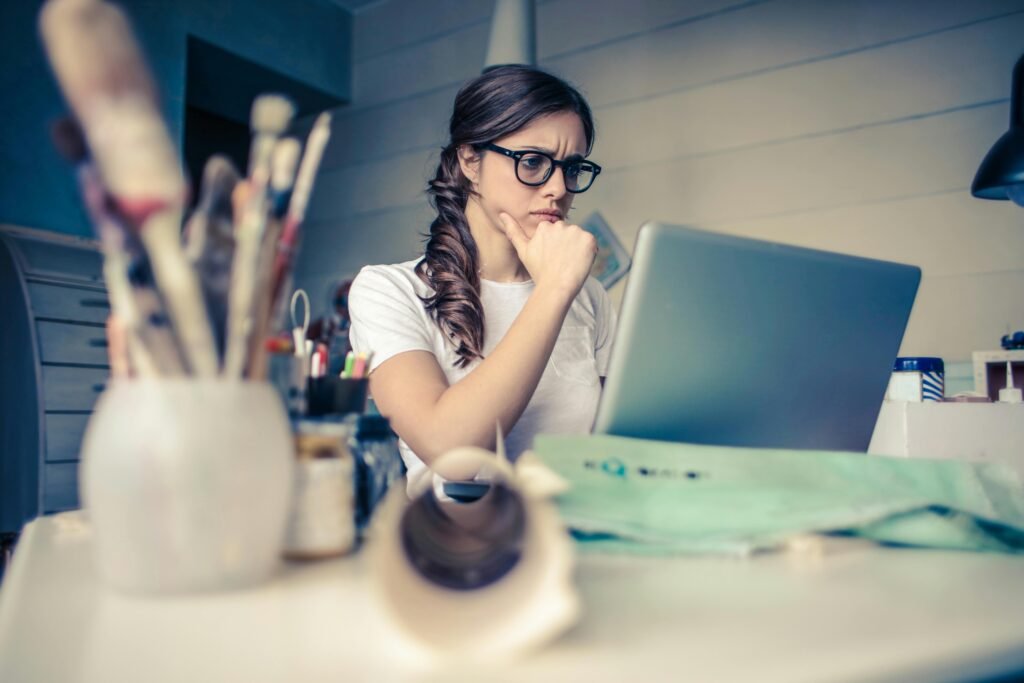 pexels-photo-941555-941555 Young woman with glasses deeply focused on a laptop surrounded by art supplies in a home office.